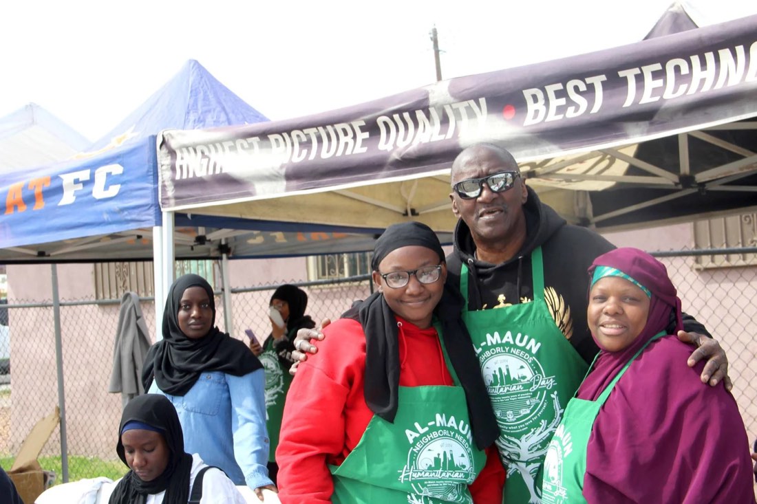 An older volunteer in a green Al-Maun apron with his arms around three smiling women, two of them also in Al-Maun aprons, under the tents at Humanitarian Day.