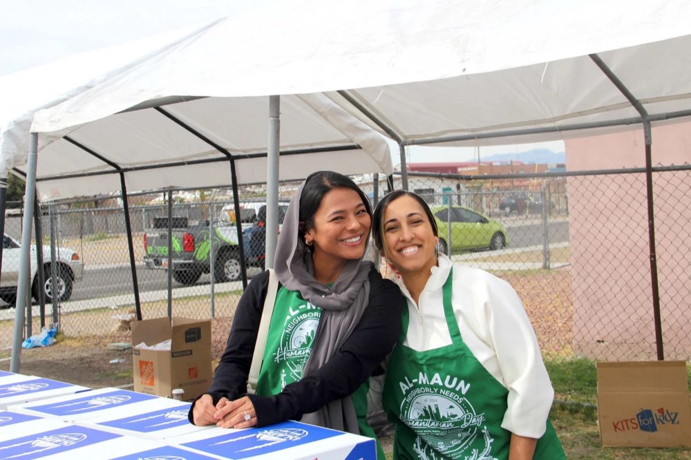 Two women volunteers in green Al-Maun aprons smiling side by side at a booth under a tent.