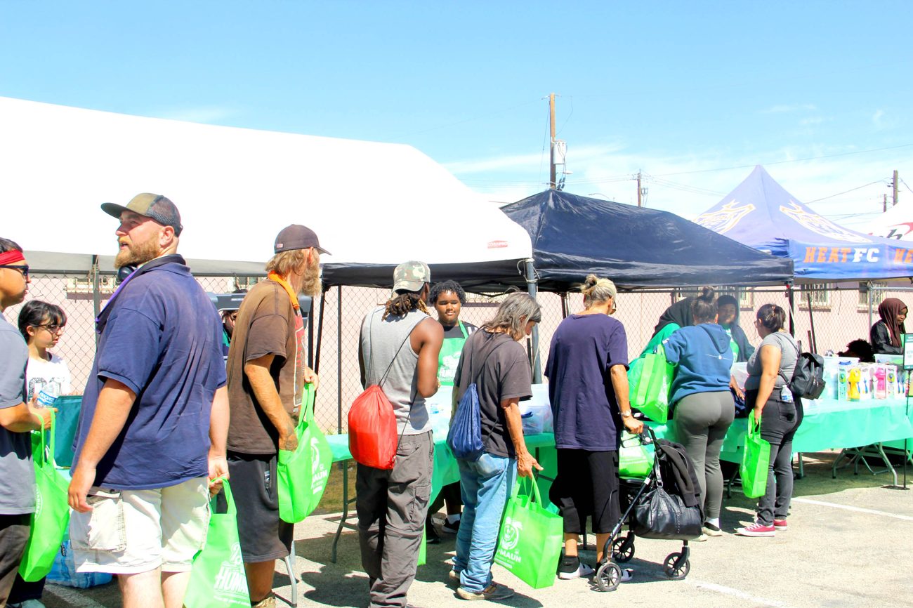 Neighbors carrying green Al-Maun bags move along the supply tables under the tents at Humanitarian Day, while volunteers in green aprons help them.