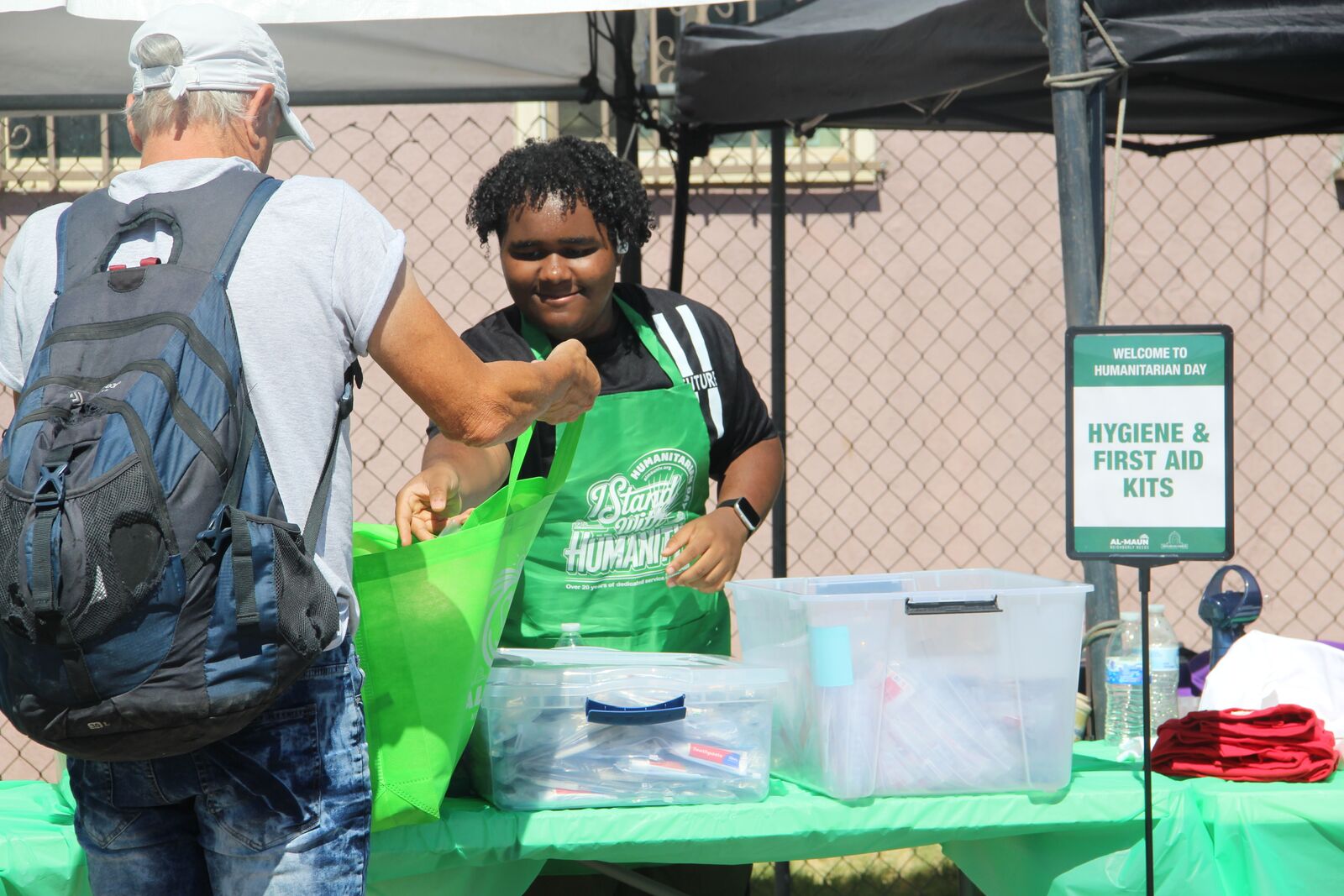 A smiling Al-Maun volunteer in a green apron handing out supplies at the hygiene and first-aid kits table.