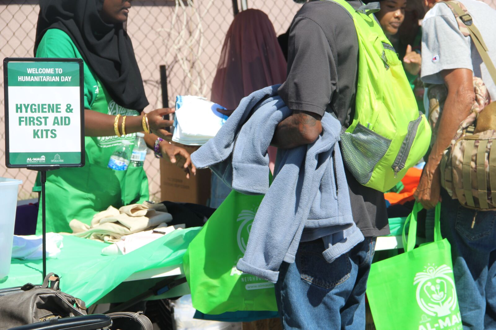 Al-Maun volunteers in green at the Humanitarian Day hygiene and first-aid kits table, with neighbors carrying green Al-Maun bags.