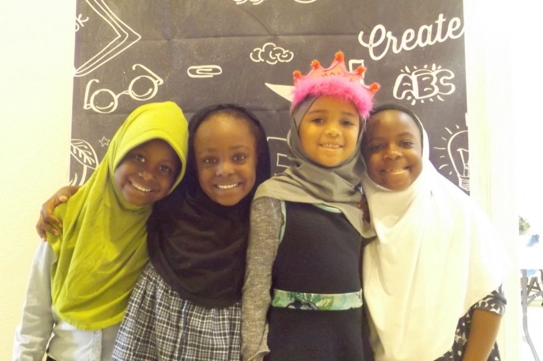 Four Nisaa&rsquo;s Academy girls laughing together, one wearing a paper crown, at a decorated &lsquo;Create&rsquo; chalkboard.