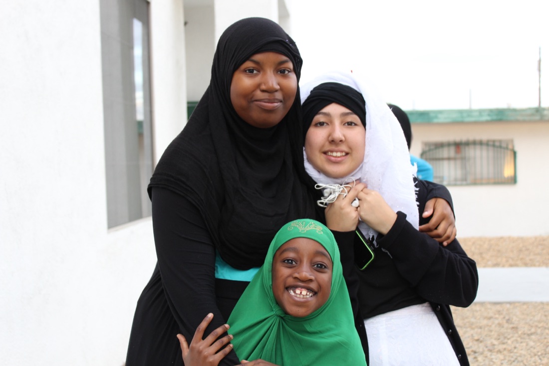 A teacher and older student embracing a smiling young girl who is waving to the camera.