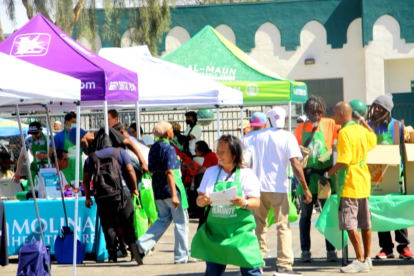 Neighbors and volunteers in green Al-Maun aprons at Humanitarian Day, with the Masjid As-Sabur arches behind.