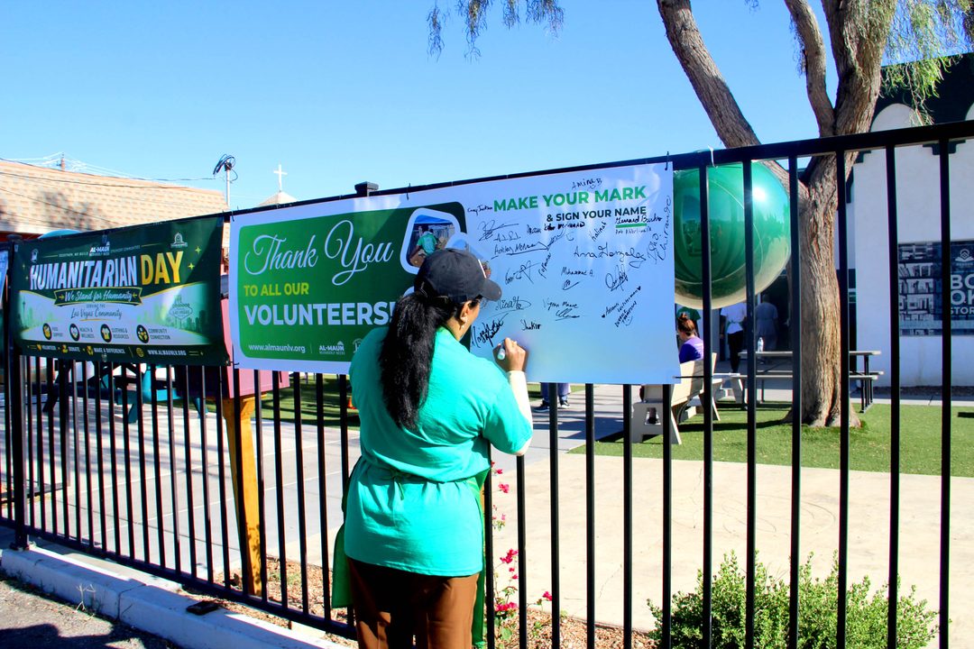 A volunteer in a green shirt signs a &ldquo;Thank you to all our volunteers&rdquo; banner hung on a fence beside a Humanitarian Day banner.