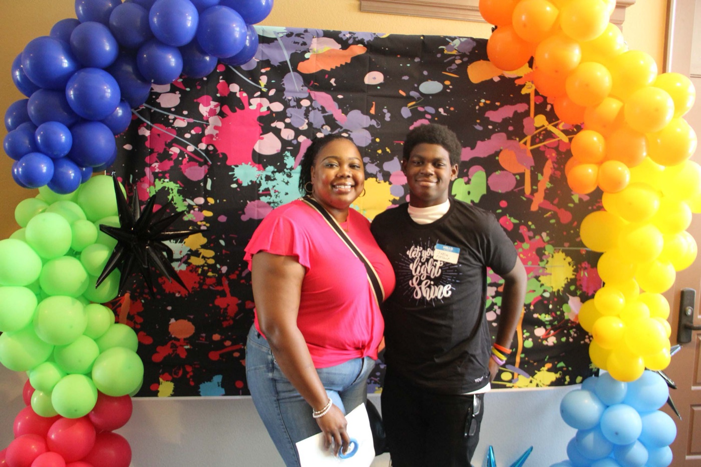 A mother and her teenage son smiling together at Al-Maun&rsquo;s Youth Conference, in front of a bright rainbow balloon arch.